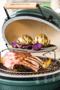 Ein geöffneter Big Green Egg Kamado-Grill mit einem gegrillten Tomahawk-Steak, gerösteten Artischocken, lila Süßkartoffeln und gegrilltem Mais.