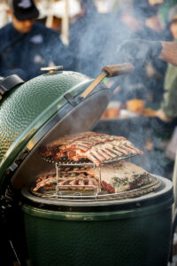 Big Green Egg mit mehreren Racks Lammkarree auf einem mehrstufigen Rosterhöhungssystem, Rauch steigt auf.