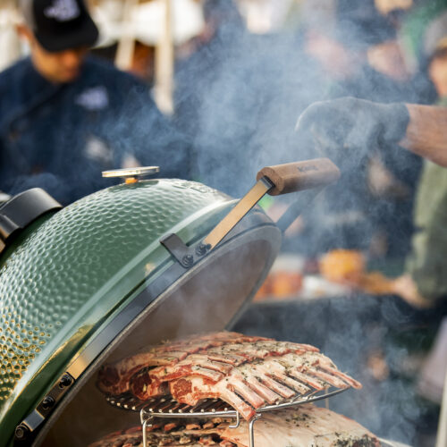 Big Green Egg mit mehreren Racks Lammkarree auf einem mehrstufigen Rosterhöhungssystem, Rauch steigt auf.