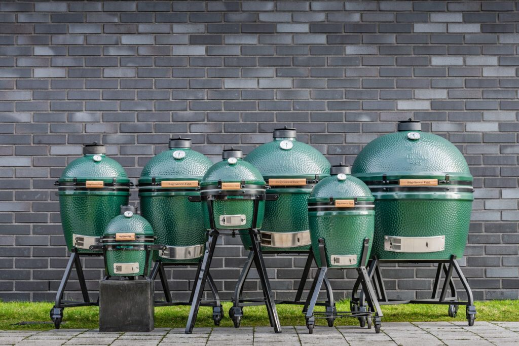 Alle Modelle des Big Green Egg Kamado-Grills in einer Reihe vor einer grauen Backsteinwand.<br />
