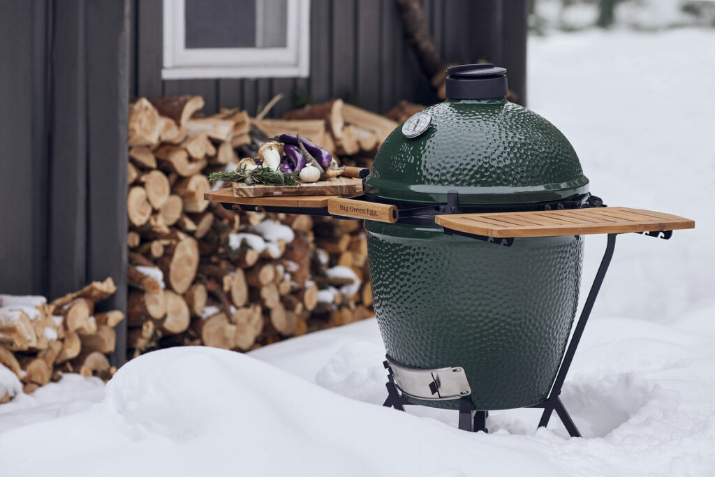 Ein Big Green Egg Large mit ausgeklappten Holzablagen, darauf frische Zutaten, im verschneiten Außenbereich vor einem Holzstapel.<br />
