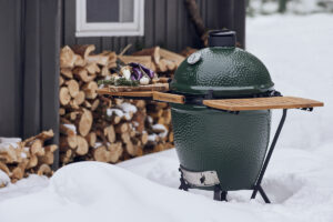 Ein Big Green Egg Large mit ausgeklappten Holzablagen, darauf frische Zutaten, im verschneiten Außenbereich vor einem Holzstapel.