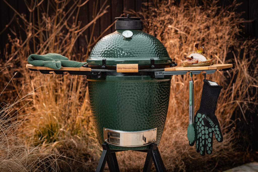 Ein Big Green Egg Medium mit Holzablagen, Grillhandschuh, Zange und einem frisch gegrillten Sandwich in herbstlicher Umgebung.<br />
