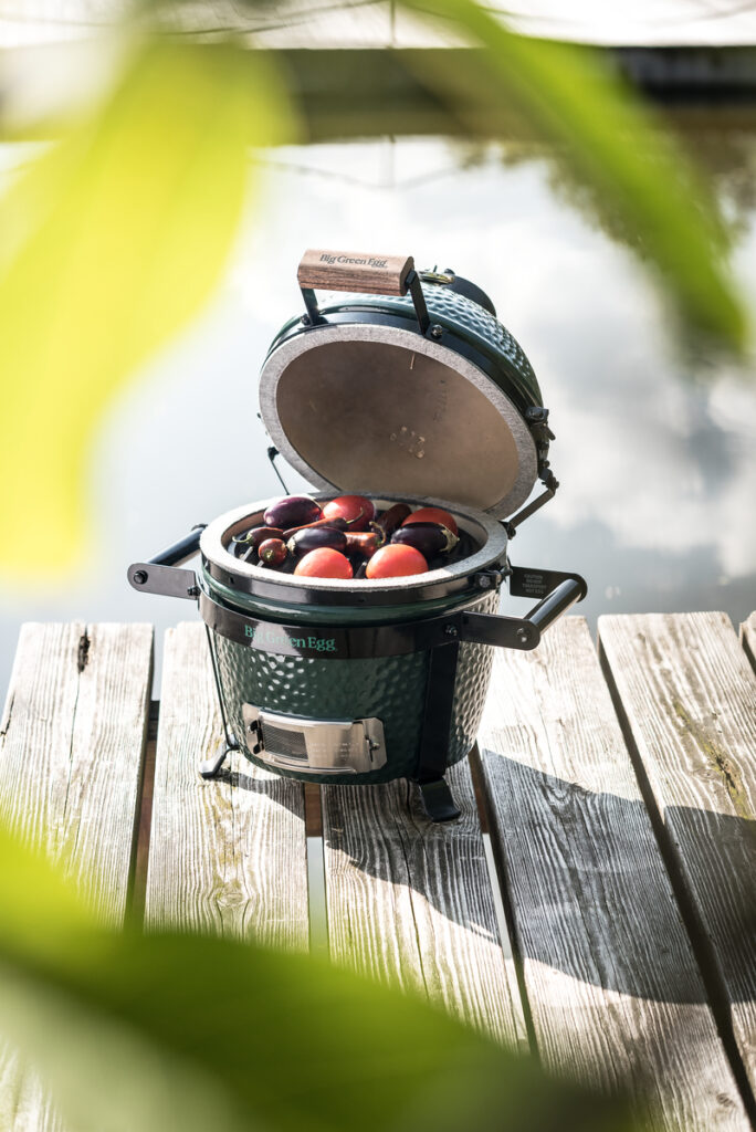 Ein Big Green Egg Mini steht geöffnet auf einem Holzsteg am Wasser und ist mit frischen Auberginen und Tomaten gefüllt.<br />
