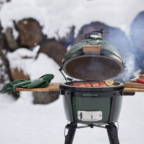 Ein Big Green Egg MiniMax steht im Schnee und grillt ein saftiges Lachsfilet mit Zitrone, während Rauch aufsteigt.