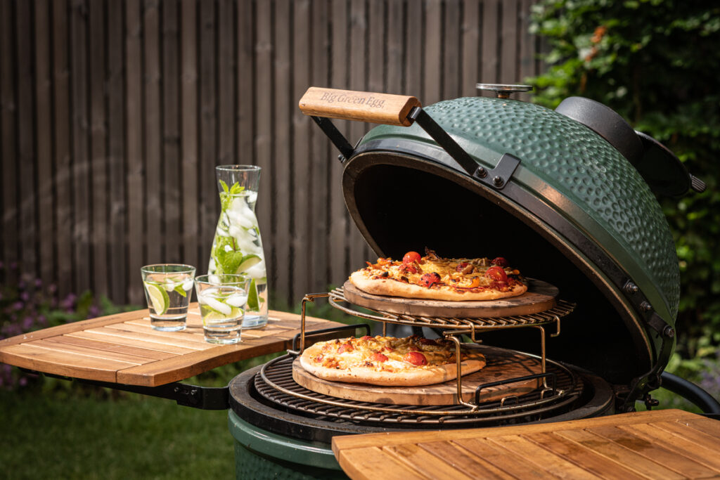 Geöffneter Big Green Egg Kamado-Grill mit zwei knusprigen Pizzen auf Pizzasteinen, daneben eine Karaffe mit Minz-Zitronenwasser.