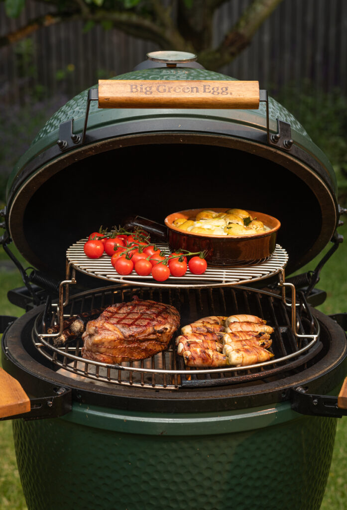 Geöffneter Big Green Egg Grill mit gegrilltem Entrecôte, Speck umwickeltem Gemüse, Kirschtomaten und Kartoffelgratin in einer Keramikform.