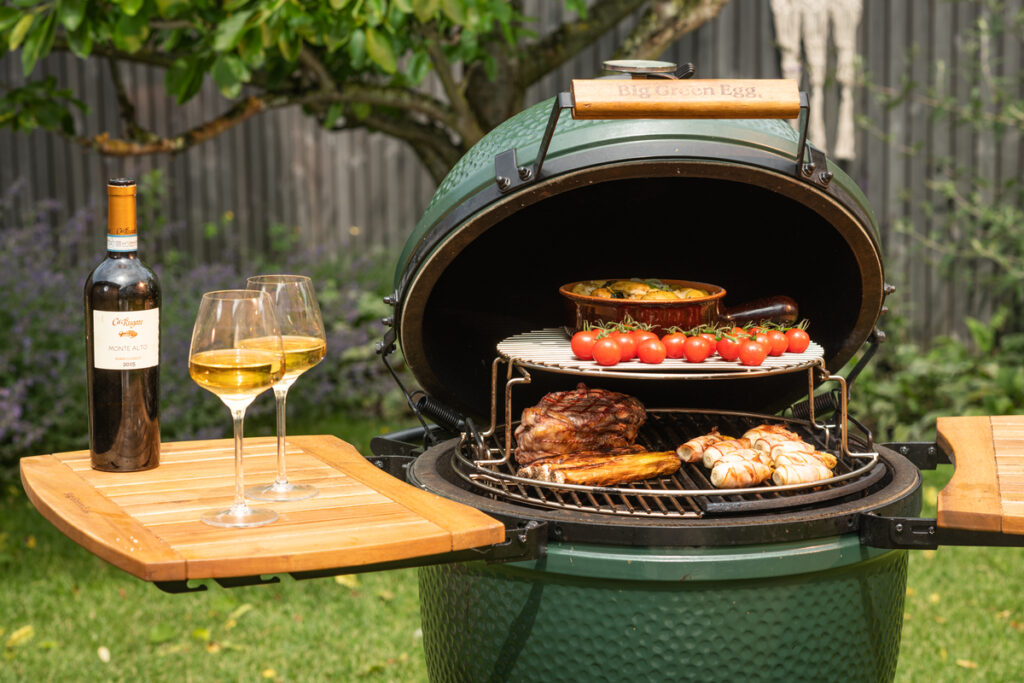 Ein geöffneter Big Green Egg Grill mit gegrilltem Entrecôte, Kirschtomaten, Kartoffelgratin und Speckröllchen, daneben eine Flasche Weißwein und zwei Gläser.<br />
