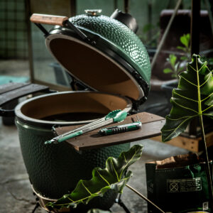 Geöffneter Big Green Egg Kamado-Grill mit Grillzange und Cutter-Messer auf der Seitenablage, umgeben von großen grünen Blättern.