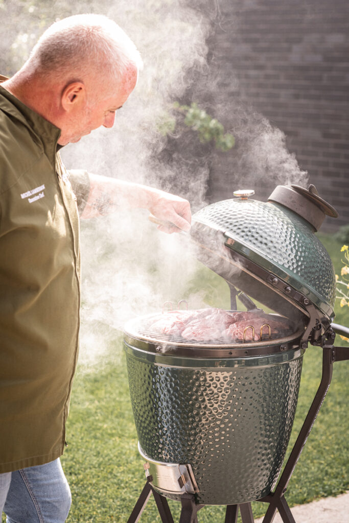 Ein Grillmeister in grüner Kochjacke öffnet den rauchenden Big Green Egg Kamado-Grill, während auf dem Rost saftiges Fleisch gegart wird.<br />
