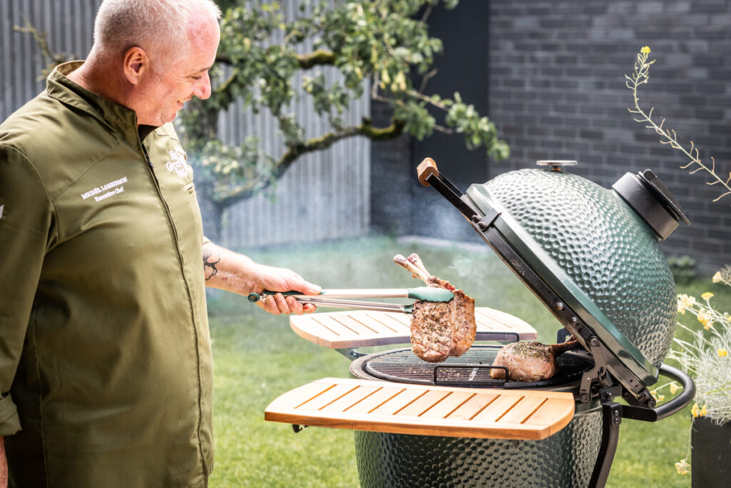 Grillmeister hebt ein gegrilltes Tomahawk-Steak mit Grillzange vom Big Green Egg<br />
