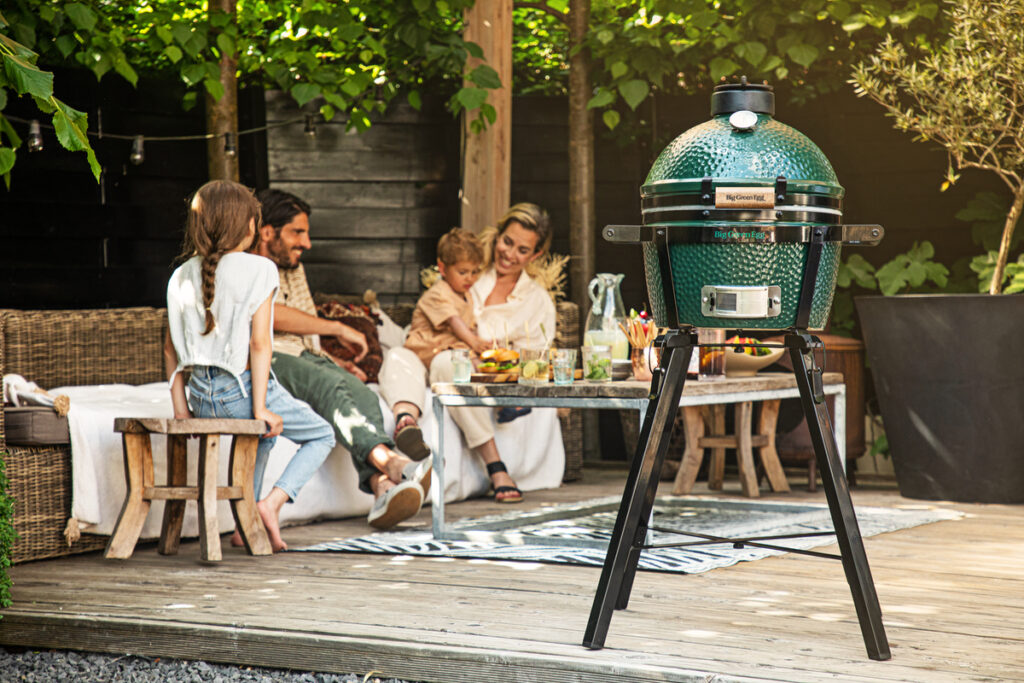 Ein Big Green Egg MiniMax Grill steht auf einer Terrasse, während eine Familie im Hintergrund gemeinsam auf einer Sitzgruppe isst und lacht.<br />
