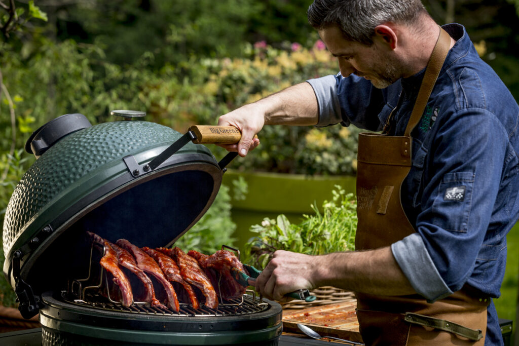 Ein Grillmeister mit Lederschürze öffnet den Big Green Egg Kamado-Grill und überprüft die langsam gegarten Spareribs.<br />
